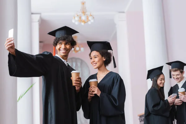 Sonriendo afroamericanos graduados tomando selfie y sosteniendo vasos de papel - foto de stock