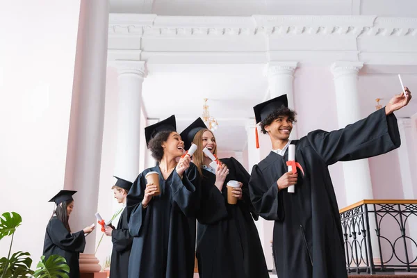 Interracial graduates with diplomas and coffee to go taking selfie in university — Stock Photo