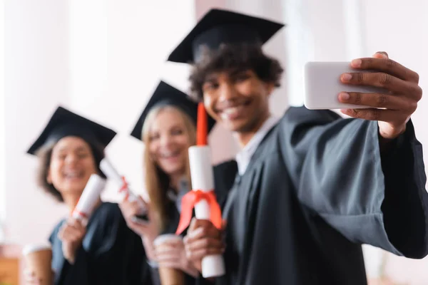 Blurred african american graduate taking selfie with smartphone near friends — Stock Photo