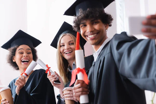 Cheerful interracial graduates with paper cups and diplomas taking selfie on smartphone — Stock Photo