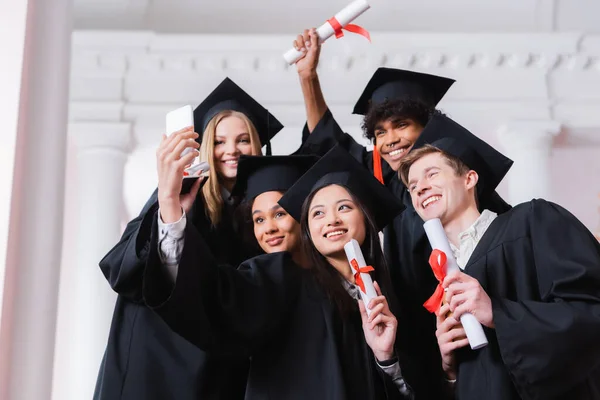 Interracial graduates with diplomas taking selfie in university — Stock Photo