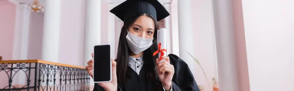 Asiático graduado en máscara médica celebración de teléfono inteligente con pantalla en blanco y diploma, bandera - foto de stock