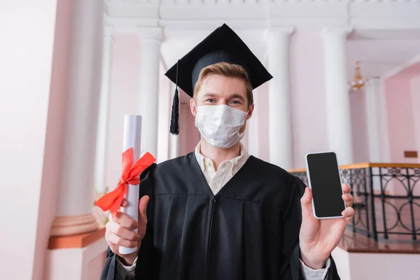 Diplômé en masque médical tenant smartphone et diplôme — Photo de stock