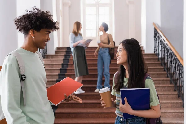 Sorrindo estudante afro-americano com notebook falando com amigo asiático com café na universidade — Fotografia de Stock