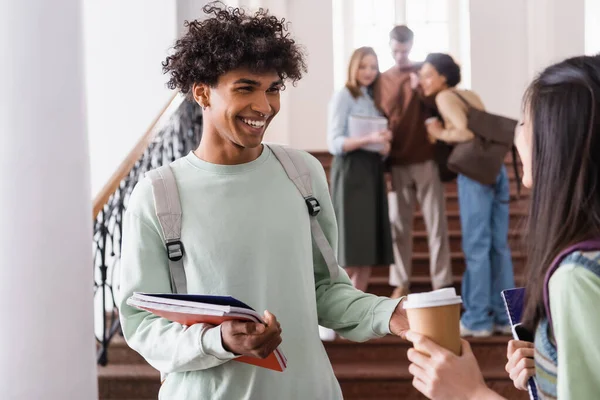 Lächelnder afrikanisch-amerikanischer Student mit Notizbüchern im Gespräch mit asiatischem Freund mit Coffee to go — Stockfoto