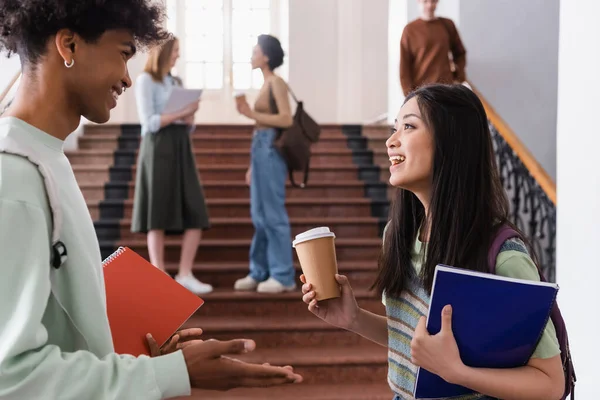 Asian student with takeaway drink talking with african american friend with notebook — Stock Photo