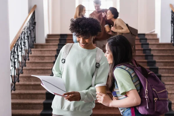 Positive african american student holding notebook near asian friend with coffee to go and backpack — Stock Photo