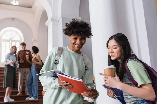 Estudiantes interracial sonrientes con mochilas y taza de papel mirando el cuaderno - foto de stock