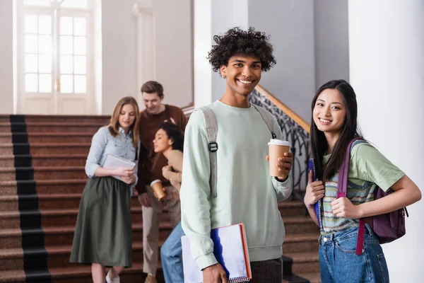 Multiethnic students with backpacks and coffee to go smiling in university — Stock Photo