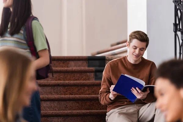 Estudiante sonriente mirando cuaderno cerca de la gente en la universidad - foto de stock