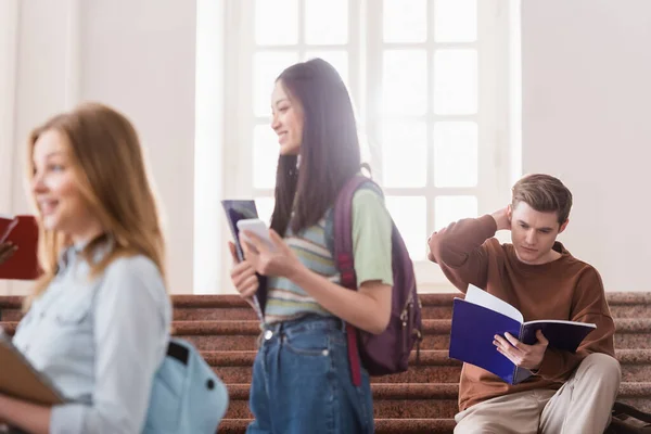 Estudiante mirando cuaderno cerca de personas multiétnicas en primer plano borrosa - foto de stock