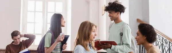 Sonriente estudiante asiático con teléfono inteligente y portátil hablando con un amigo afroamericano, bandera - foto de stock