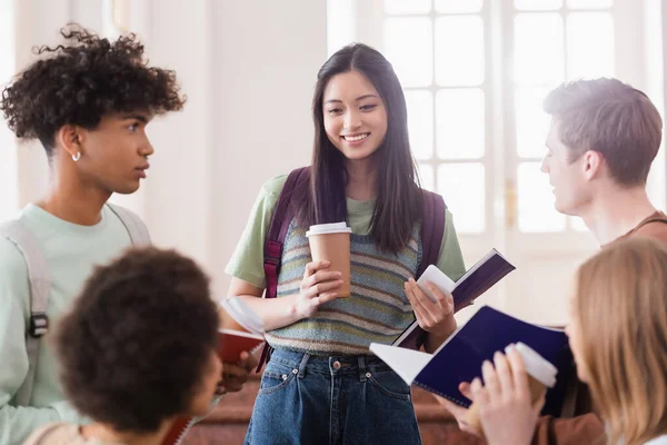 Sonriente estudiante asiático con café y teléfono inteligente de pie cerca de amigos multiétnicos - foto de stock