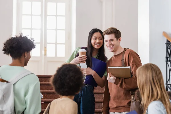 Étudiants interracial souriants avec café et cahiers regardant des amis flous — Photo de stock