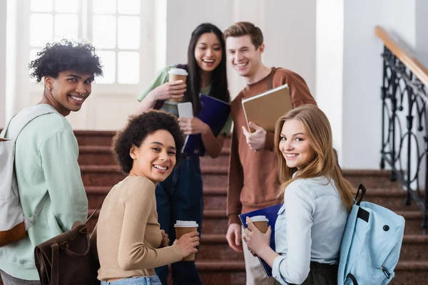 Alegre interracial estudiantes con café para ir y mochilas mirando a la cámara - foto de stock