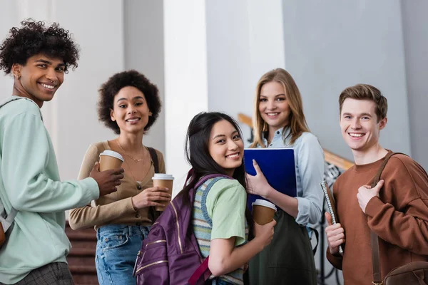 Estudiantes multiétnicos con cuadernos y papeles tazas sonriendo a la cámara - foto de stock