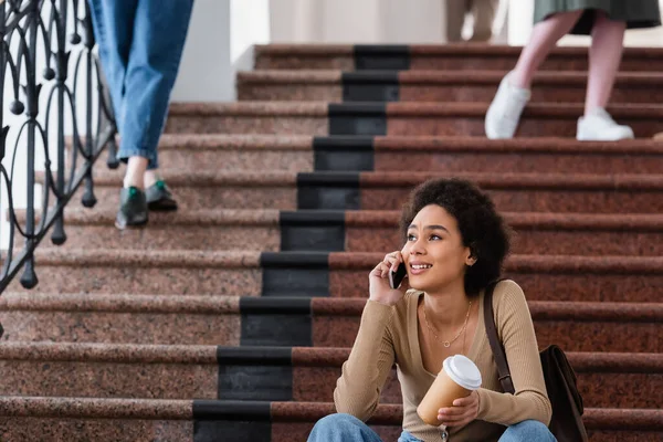 Усміхаючись афроамериканською студенткою з кавою, розмовляючи на смартфоні в університеті. — стокове фото