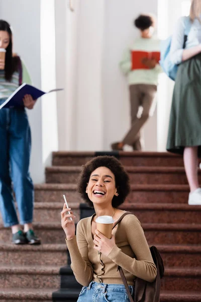 Cheerful african american student holding paper cup and smartphone in university — Stock Photo