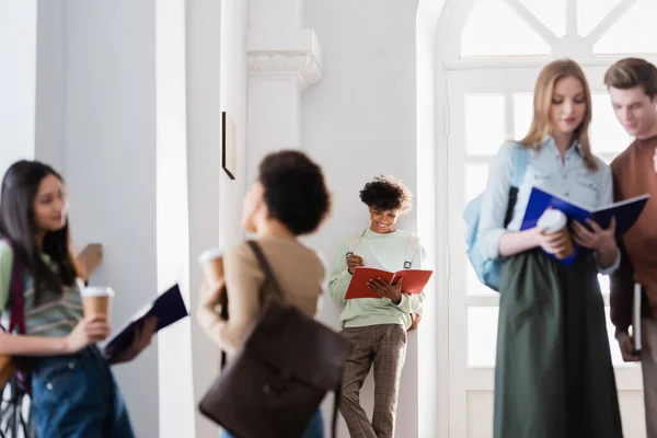 Lächelnder afrikanisch-amerikanischer Student schaut auf Notizbuch in der Nähe verschwommener Menschen an der Universität — Stockfoto