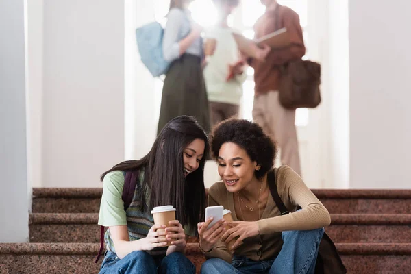 Sorrindo estudantes multiétnicos com café para ir usando celular — Fotografia de Stock