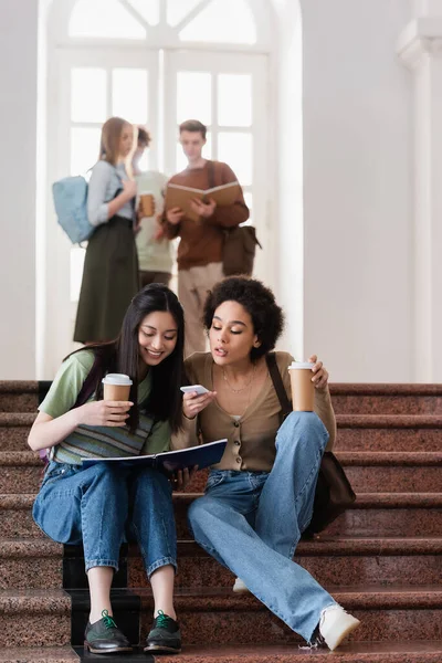 Interrassische Studenten mit Coffee to go, Notebook und Smartphone sitzen auf der Treppe — Stockfoto