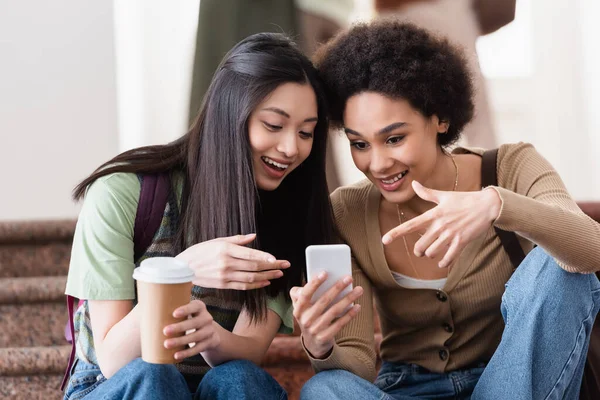 Smiling multiethnic students with coffee to go using mobile phone — Stock Photo