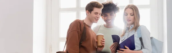 Interracial students with coffee to go and notebook standing near window, banner — Stock Photo