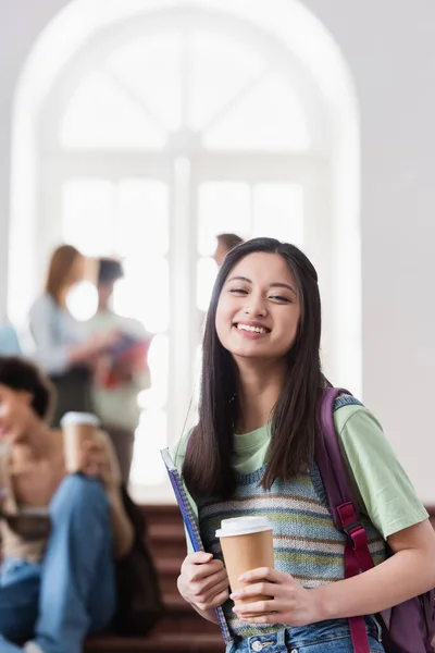 Sonriendo asiática estudiante con papel taza y portátil mirando a la cámara - foto de stock