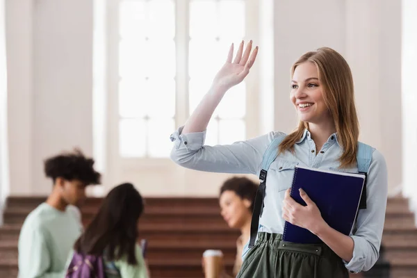 Glückliche Studentin mit winkendem Notizbuch in Uni-Aula — Stockfoto