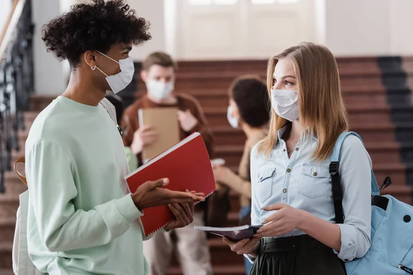 Studenti interrazziali in maschere mediche che tengono quaderni e parlano — Foto stock