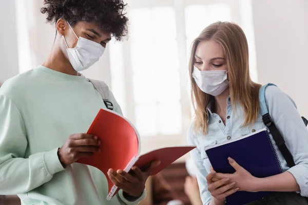 Estudiante afroamericano en máscara médica sosteniendo cuaderno cerca de amigo - foto de stock