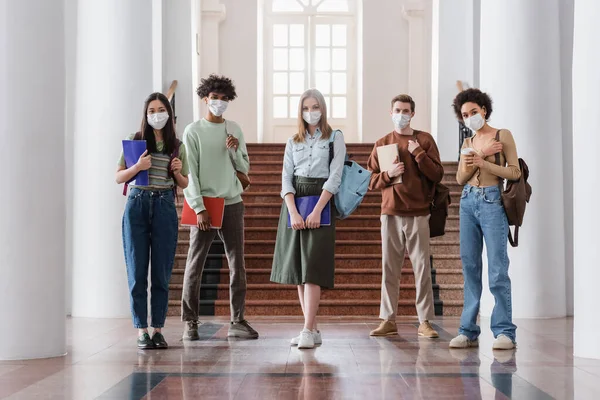 Interracial students in medical masks holding notebooks in university — Stock Photo