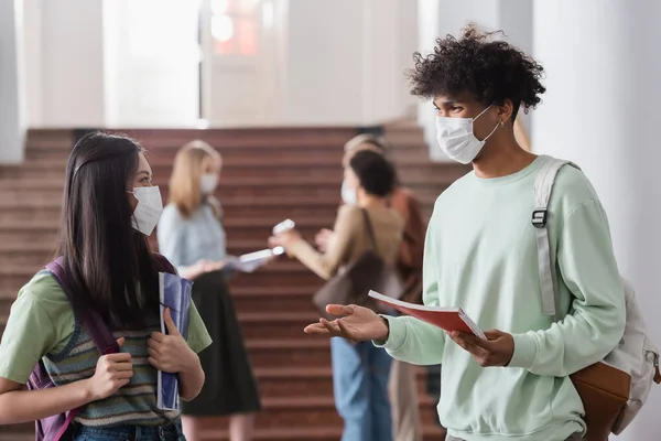 Estudiantes multiétnicos con máscaras médicas sosteniendo cuadernos y hablando - foto de stock