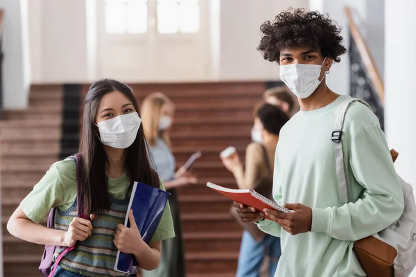 Estudiantes asiáticos y afroamericanos en máscaras médicas sosteniendo cuadernos - foto de stock