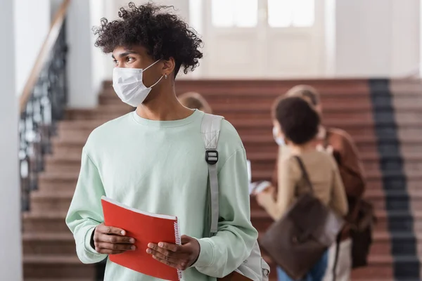 Estudiante afroamericano en máscara médica sosteniendo cuaderno en la universidad - foto de stock
