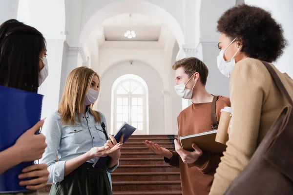 Estudiantes en máscaras médicas apuntando con las manos cerca de amigos multiétnicos con cuaderno y café - foto de stock