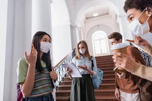 Asian student in medical mask talking to african american friend with coffee on blurred foreground — Stock Photo