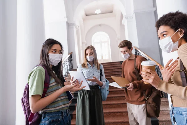 Studenti interrazziali con quaderni e coppa di carta che parlano nella sala dell'università durante la quarantena — Foto stock