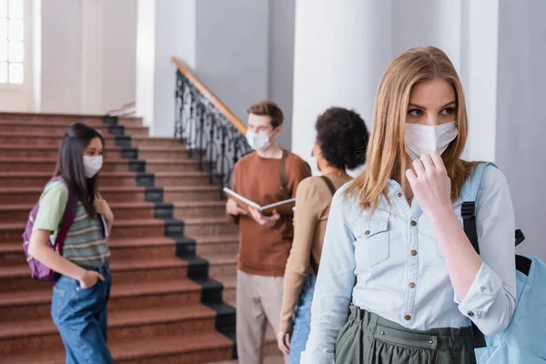 Student in medical mask standing near blurred multiethnic friends in university — Stock Photo