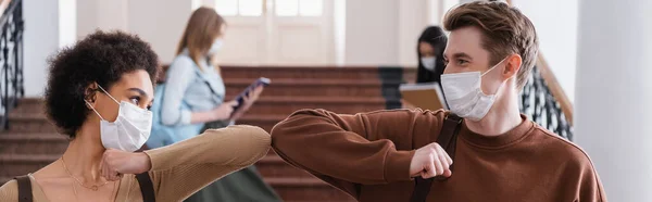 Estudiantes interraciales con máscaras médicas haciendo golpes de codo, pancarta - foto de stock