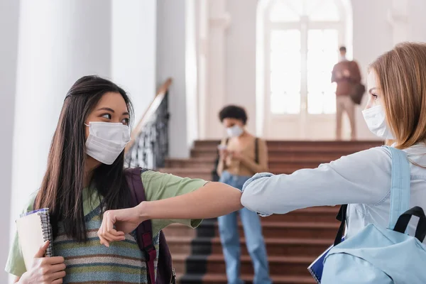 Estudiantes multiétnicos en máscaras médicas saludan con el codo golpeado - foto de stock