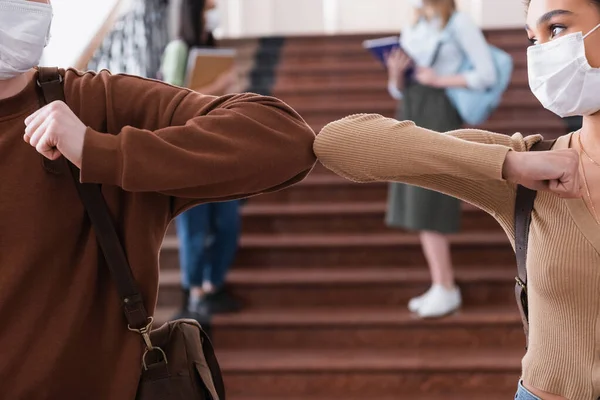 Interracial students in medical masks doing fist bump in university hall — Stock Photo