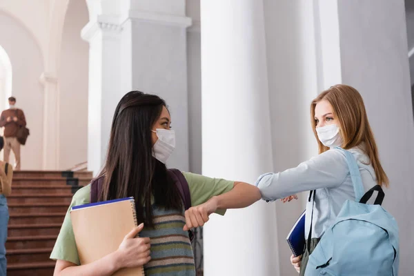 Estudiantes multiétnicos con máscaras protectoras sosteniendo cuadernos y saludos en el salón universitario - foto de stock