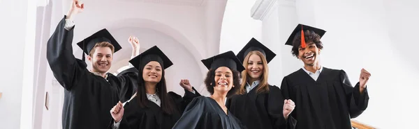 Cheerful multiethnic students in academic gowns showing yes gesture, banner — Stock Photo
