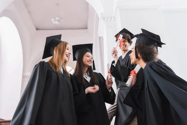 Vue en angle bas des diplômés souriants interracial avec des diplômes dans la salle universitaire — Photo de stock