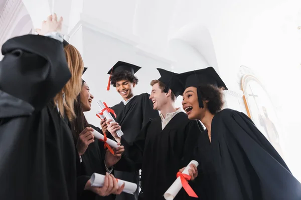Vista de ángulo bajo de los graduados multiétnicos felices con diplomas en el salón de la universidad - foto de stock