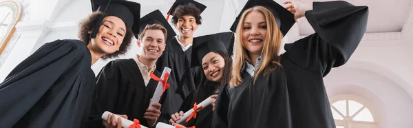 Low angle view of smiling interracial graduates holding diplomas, banner — Stock Photo