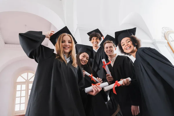 Vista de ángulo bajo de los graduados multiétnicos con diplomas sonriendo a la cámara - foto de stock