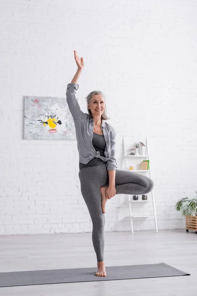 Mujer de mediana edad feliz en ropa deportiva practicando yoga en la estera en casa - foto de stock
