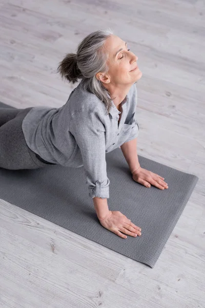 Pleased middle aged woman in cobra pose practicing yoga on mat at home — Stock Photo